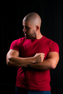 Retrato de homem careca jovem em est&uacute;dio com fundo preto. homem vestindo blusa vermelha com bra&ccedil;os cruzados, olhando para o bra&ccedil;o musculoso.