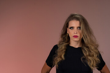 Fototapeta premium Retrato de mulher loira de olhos verdes, cabelo cacheado, roupa preta em fundo cor salmão no estúdio. Jovem olhando para a câmera de forma séria.