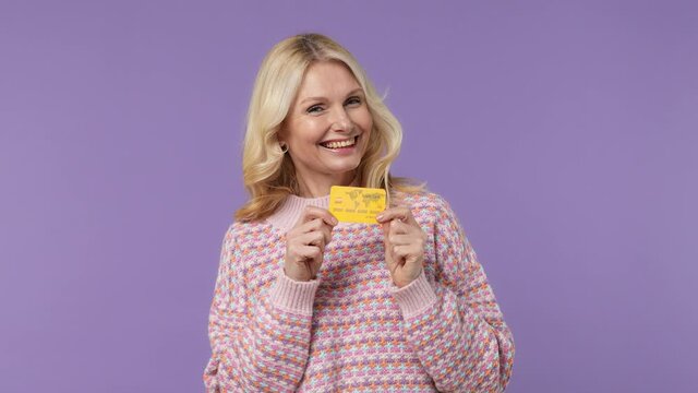 Satisfied elderly blonde woman lady 40s years old in warm shirt point index finger on mockup plastic credit bank card showing thumbs up isolated on plain pastel light purple background studio portrait
