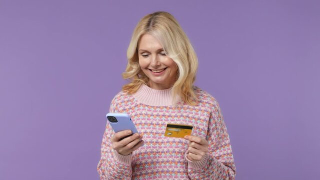 Dreamful elderly blonde woman lady 40s years old in warm shirt using mobile cell phone hold in hand credit bank card do online shopping isolated on plain pastel light purple background studio portrait