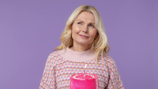 Excited fun surprised elderly blonde woman lady 40s years old wears warm shirt cover hide face with hands birthday cake blow out candle isolated on plain pastel light purple background studio portrait