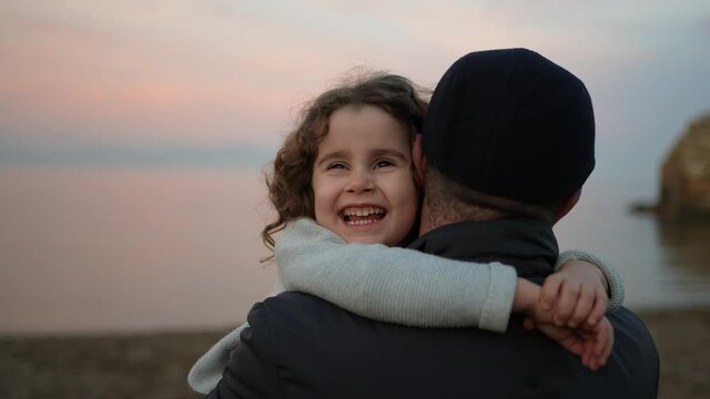 Portrait of a pretty little girl smiling and hugging her father, grandfather at sunrise on the beach. Family happiness concept