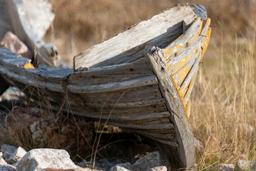 A wooden boat, painted yellow, damaged by time and weather. The open rowboat has the back rotted and is missing boards. It is among tall yellow grass and large rocks on a shoreline. 