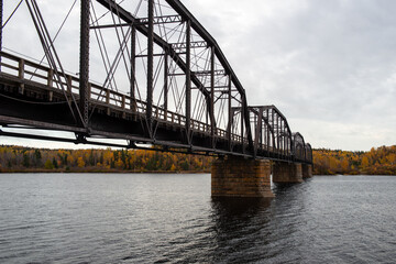 Steel trestle bridge with wooden deck over a large river in Newfoundland.  The bridge is for foot traffic and ATV usage. The sky is clear blue and land and houses can be seen in the background. 