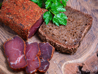 Top view of sliced slices of basturma (dried meat) with parsley and a piece of rye bread on a wooden background. Rustic homemade food. Juicy pieces of meat