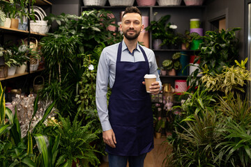 florist entrepreneur in his shop surrounded by flowers and potted plants stands with a cup of coffee and looks with a smile at the camera.