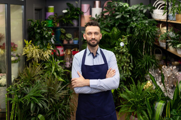 a florist man in a boutique of bouquets and flowers stands with a smile on his face in a blue apron with his arms crossed on his chest.