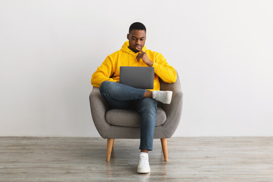 Black Man Using Laptop Sitting In Armchair Near Gray Wall