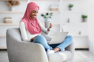 Obraz premium Happy Black Muslim Woman Relaxing With Laptop And Drinking Coffee At Home