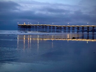 The ocean and beach at night with reflections of lights hanging on an old wooden pier glowing in the ocean water and damp sand cloudy blue skies in this ocean scene of relaxation and calmness