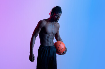 Portrait of motivated black basketballer with naked torso wearing headphones and holding ball in...