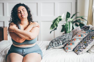 Curly haired overweight young woman in grey top and shorts with satisfaction on face accepts curvy body shape standing in stylish bedroom closeup - obrazy, fototapety, plakaty