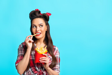 Young pinup woman in retro outfit eating french fries from package, looking aside at empty space on blue background