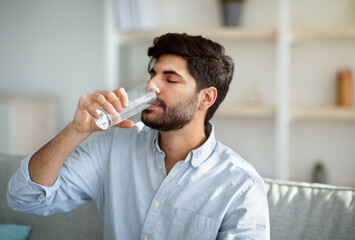 Young arab man drinking fresh water from glass, sitting on sofa in living room interior, copy space - Powered by Adobe