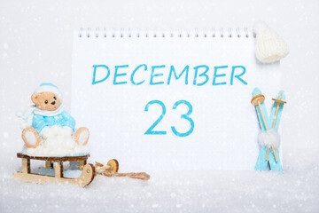 December 23rd. Teddy bear sitting on a sled, blue skis and a calendar date on white snow. Day 23 of month.
