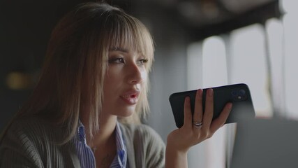 Asian woman talk on cell using voice assistant. Woman sitting on couch holds phone talks on speakerphone with friend makes voice recognition or request uses internet services through virtual assistant