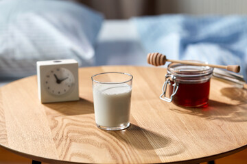 sleep disorder, bedtime and morning concept - close up of glass of milk, honey and alarm clock on night table at home