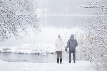 Young couple standing on snow at lake shore and holding each other hands and looking far away. Romantic lovely moment at beautiful place. Peaceful atmosphere. White cold snowy winter day. Back view. - Powered by Adobe