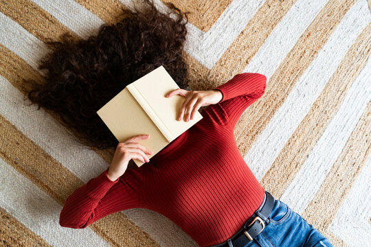 Woman covering face with book lying on carpet at home