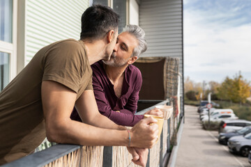 Romantic gay couple holding coffee mugs kissing in balcony