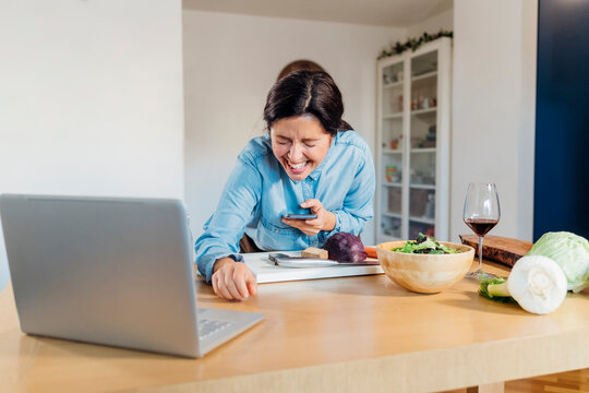 Woman holding smart phone laughing on video call using laptop at home