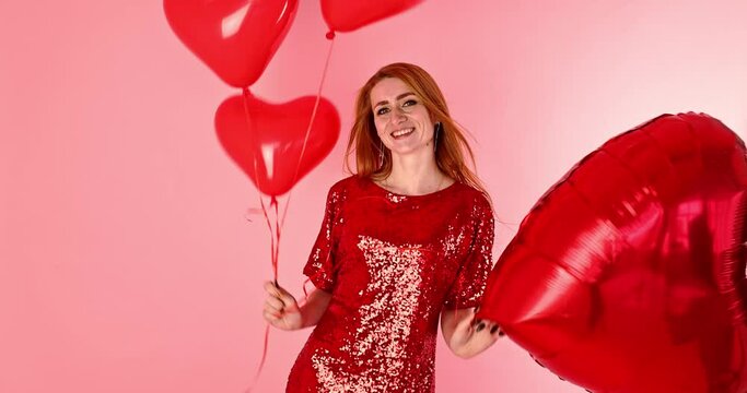 Beautiful redhead girl with red heart balloon posing. Happy Valentine's Day concept. Studio photo of beautiful ginger girl dancing on pink background.