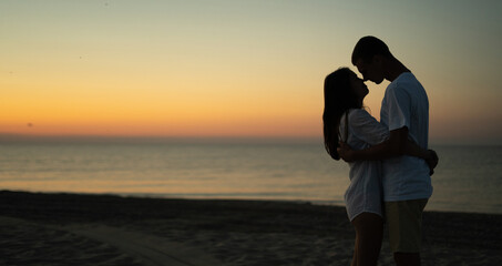 Fototapeta premium Two young lovers stand on the beach and look at each other against the backdrop of the sunset. Summer vacation concept while vacationing on a tropical beach. Baner.
