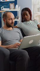 Interracial couple with pregnancy relaxing together at home. Pregnant black woman switching channels with TV remote control and caucasian man using laptop while sitting in living room