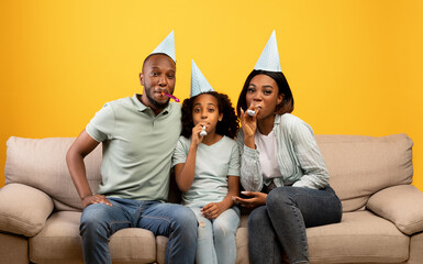 Happy african american parents and girl celebrating birthday and blowing at party horns over yellow background