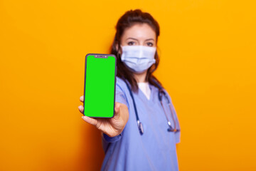 Nurse holding vertical green screen on smartphone while wearing face mask against coronavirus....