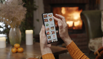 Ordering food using a smartphone at home. A woman selects sushi sets in the internet menu of a Japanese restaurant using an application on a smartphone. Home evening furnishings with a fireplace.