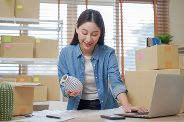 Small business entrepreneur SME, asian young woman owner packing product, checking parcel box delivery, using laptop receive order from customer, working at home office. Merchant online, ecommerce.