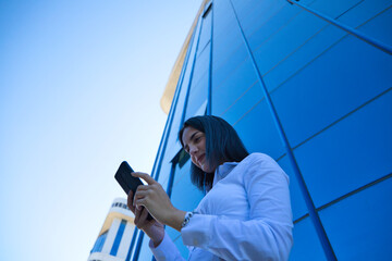 young, beautiful woman is consulting her mobile phone at the entrance door of the building where she works. The photo is taken from below and the building can be seen. Concept businesswoman.