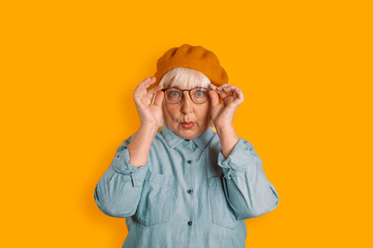 Photo Of Blonde Hair 50s Woman Astonished Face Touch Glasses Look Camera Isolated Over Orange Color Background