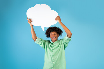 Positive african american teenager holding empty speech bubble above his head over blue background,...