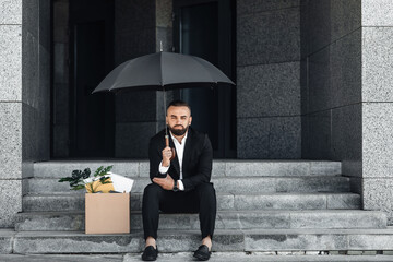 Unemployment concept. Jobless mature businessman sitting under umbrella with empty poster and box of belongings
