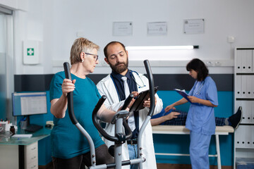 Doctor consulting old patient doing gymnastics for recovery at physiotherapy clinic. Aged woman using stationary bicycle for physical exercise and fitness activity while talking to medic