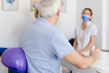 Obraz premium older man sitting with a pilates and yoga medicine ball on his back to rehabilitate from shoulder tendonitis, while talking to the occupational therapist, a physiotherapist