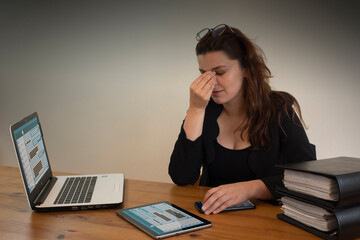 Beautiful Hispanic female office worker massaging her eyes with her left hand, sitting at her desk in front of her computer