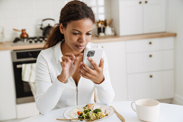 Young black woman using cellphone while having lunch at home