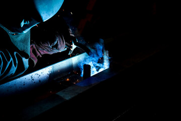 A man wearing a welding mask welds metal parts at a factory.