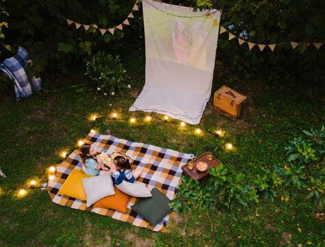 High Angle View Of Children Sitting On Plaid And Looking Movie On Backyard. Summer Outdoor Weekend Activities With Kids. Open Air Cinema.