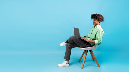 E-learning concept. African american student guy using laptop, sitting in chair on blue background, panorama, free space