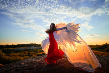 Beautiful young woman or girl in red dress and white wings on the sand on sunny day with blue sky. Angel model or dancer posing in photo shoot on dunes