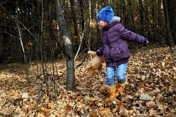 A girl child in motion holds a teddy bear in her hand. Autumn walk through the forest, dry leaves. Yellow Rubber Boots