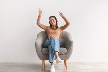 Cool young lady sitting in armchair, listening to music in wireless headphones, dancing to her favorite song
