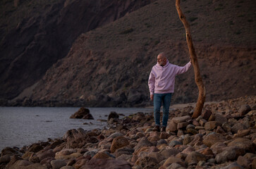 Adult man in pink standing on rocky beach with tree trunk looking at sea. Almeria, Spain