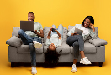 Gadgets addiction. Young black family of three using different electronic devices, sitting on sofa...