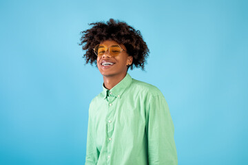 Portrait of happy african american teen guy in mint shirt and yellow sunglasses posing on blue...