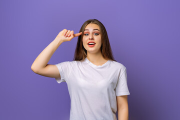 Fototapeta premium Eureka, finally got answer on question. Attractive pleasant young brunette woman thinking with finger to her temple, recalling or remembering, standing in blank white t shirt over purple background